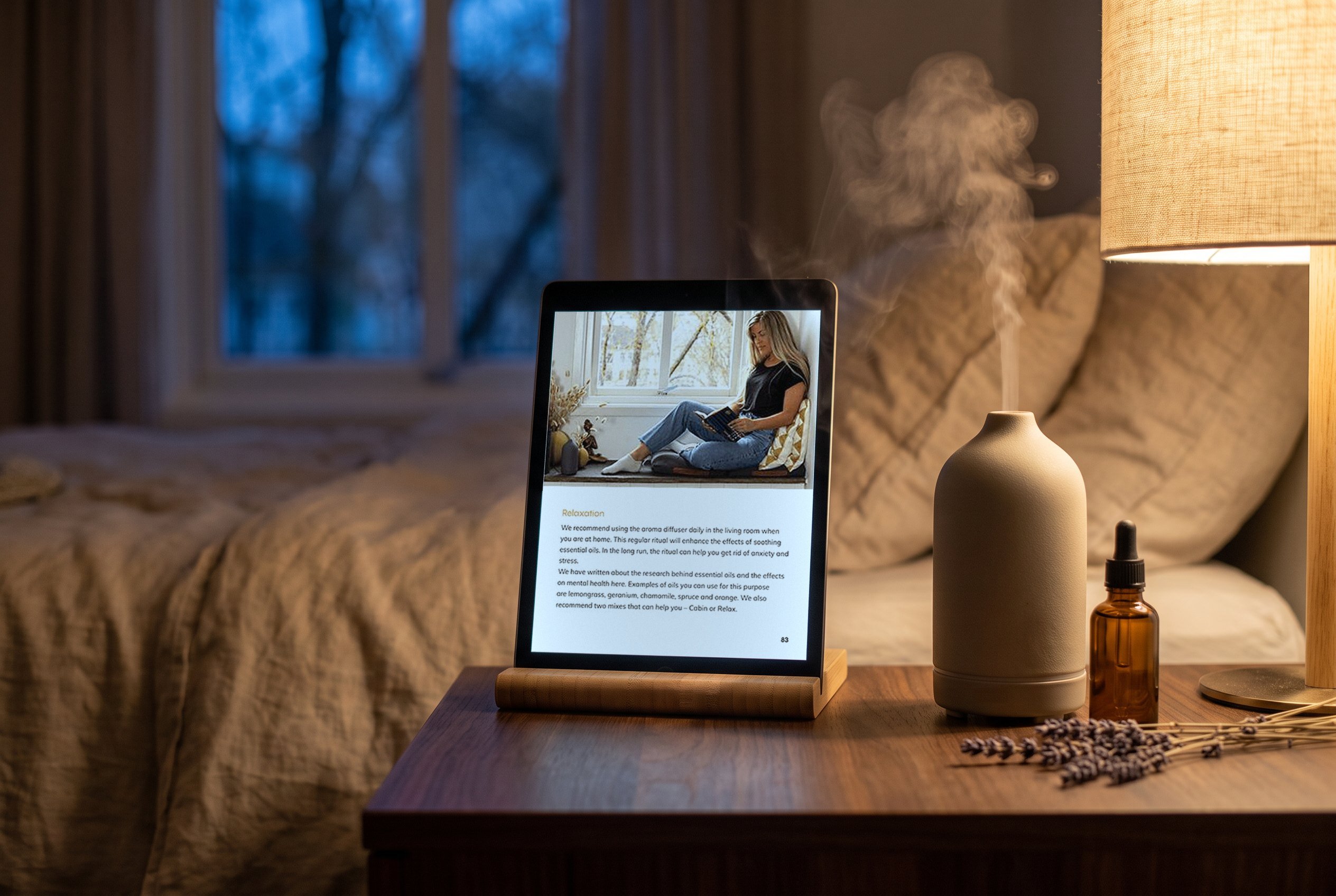 The ebook's Relaxation page on an iPad by the bed at dusk, beside a ceramic diffuser misting, a lamp and a small oil bottle