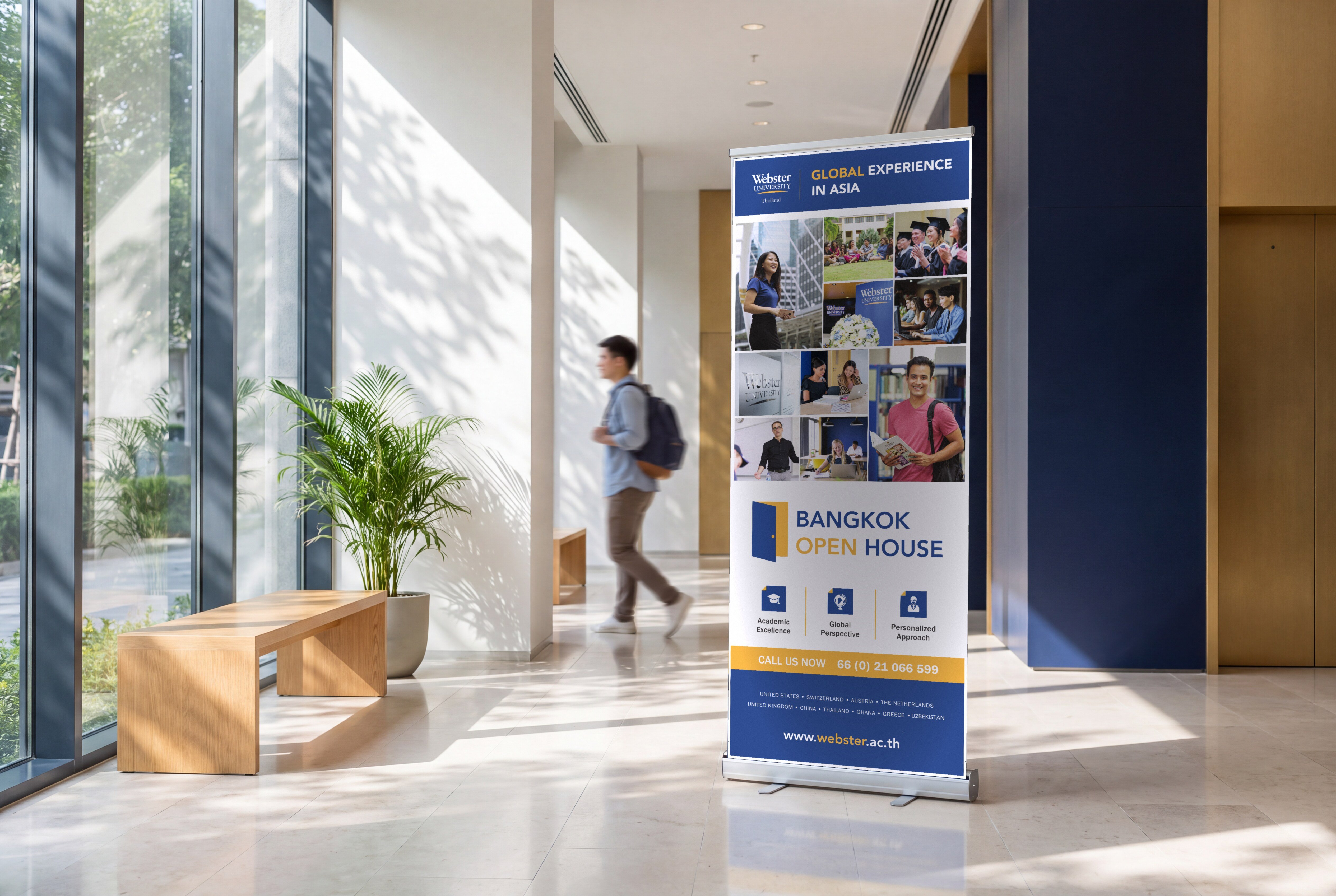 A Webster Bangkok Open House roll-up banner in navy and gold standing in a sunlit campus lobby, a student walking past